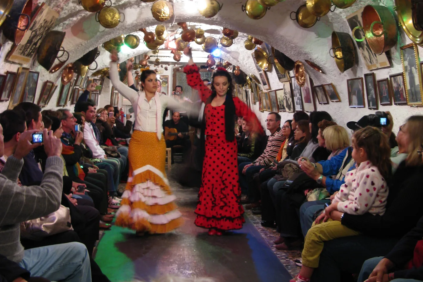 Two flamenco dancers, one in an orange skirt and white blouse and one in a red polka-dot dress, move down a narrow aisle under a whitewashed cave arch hung with copper pots, with spectators seated along both walls and a guitarist behind them